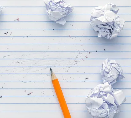A yellow pencil on a sheet of lined notepaper, with erased-out writing and several crumpled balls of paper