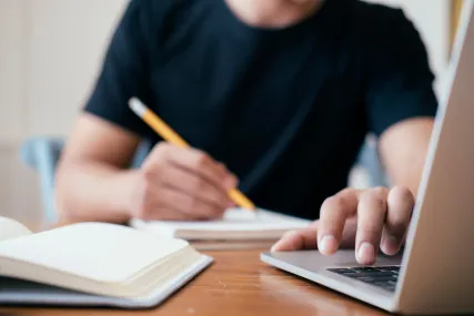 A person sits with pencil in hand at a laptop computer, scribbling and typing.