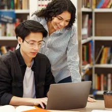 A student types on a laptop in a library while a faculty mentor leans over him.