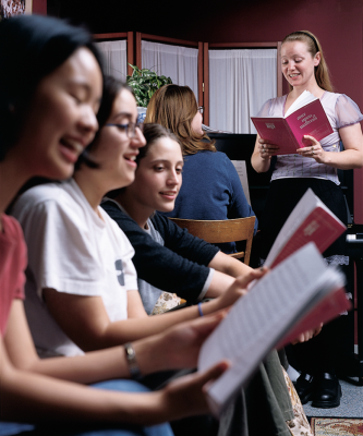 A group of women practice a vocal number as one plays the piano