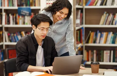 A student types on a laptop in a library while a faculty mentor leans over him.